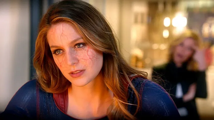 Supergirl herida Kara con el traje de Supergirl y grietas rojizas en la cara, con una mujer rubia desenfocada al fondo