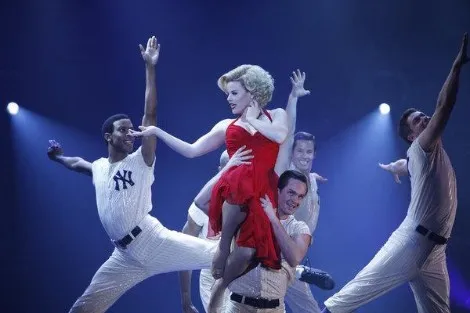 Una bailarina rubia con vestido rojo en brazos de bailarines vestidos de béisbol en un número de Smash
